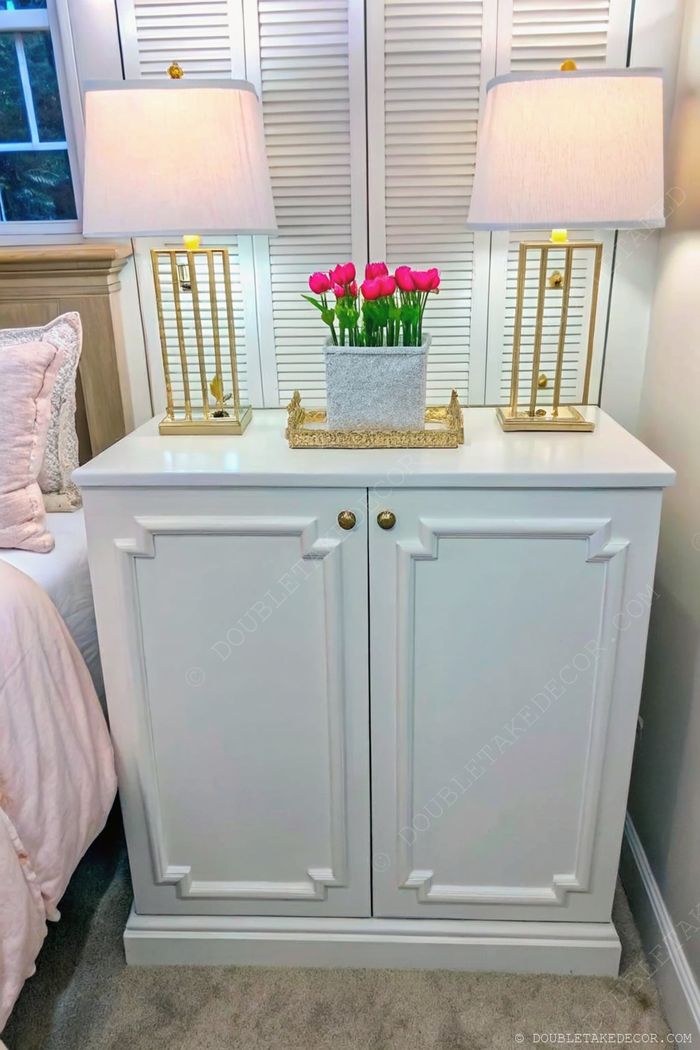 White paneled nightstand with modern gold lamps, pink tulips in a gray planter, set against wooden shutters on a soft carpet.