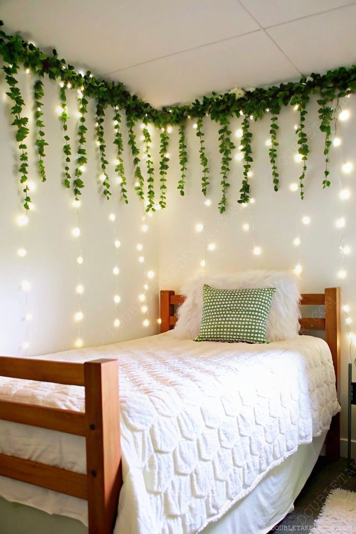 Cozy dorm bedroom with wooden bed frame, white duvet, accent pillows, and walls adorned with cascading greenery and fairy lights.