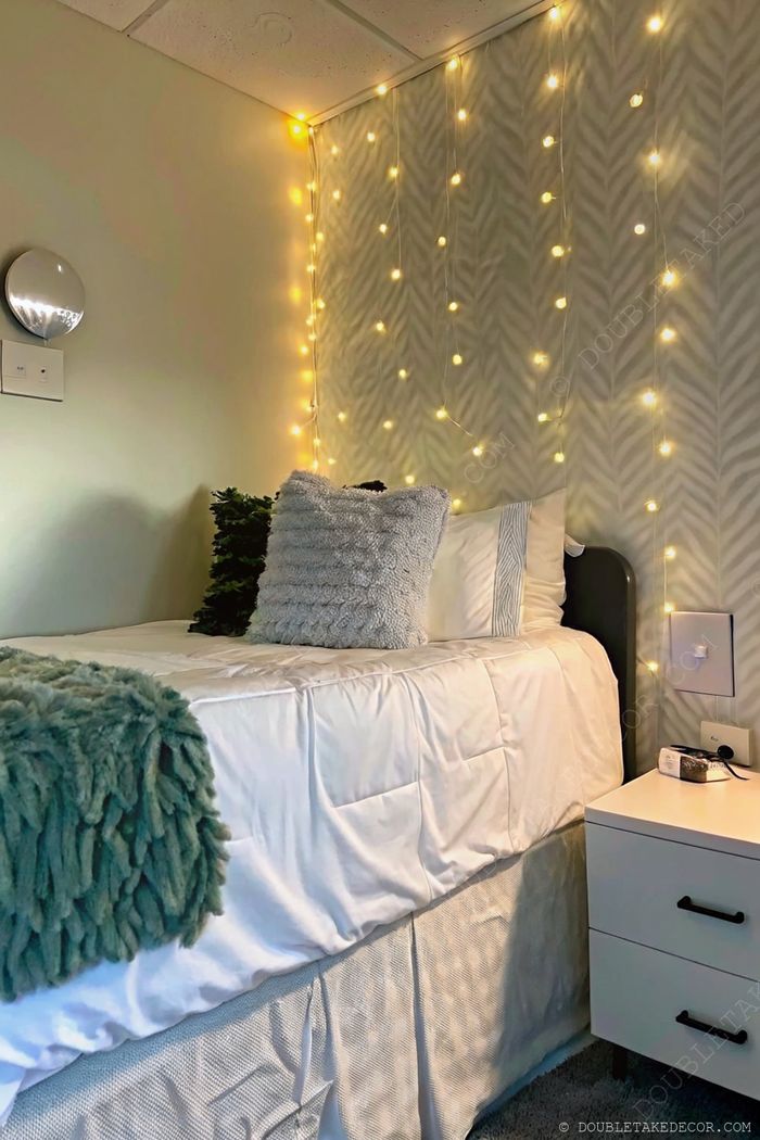 Cozy dorm bedroom corner with white bedding, green accents, twinkling fairy lights, and a stylish bedside table on gray carpet.