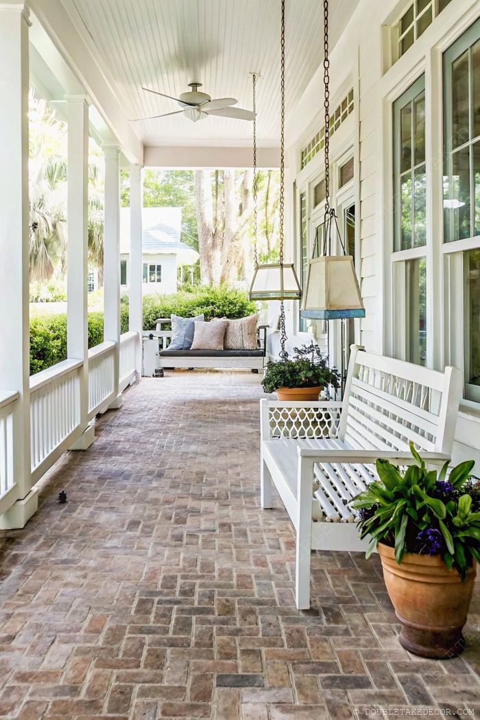 Inviting front porch with white columns, ceiling fan, decorative light, brick flooring, and a comfortable swing with cushions