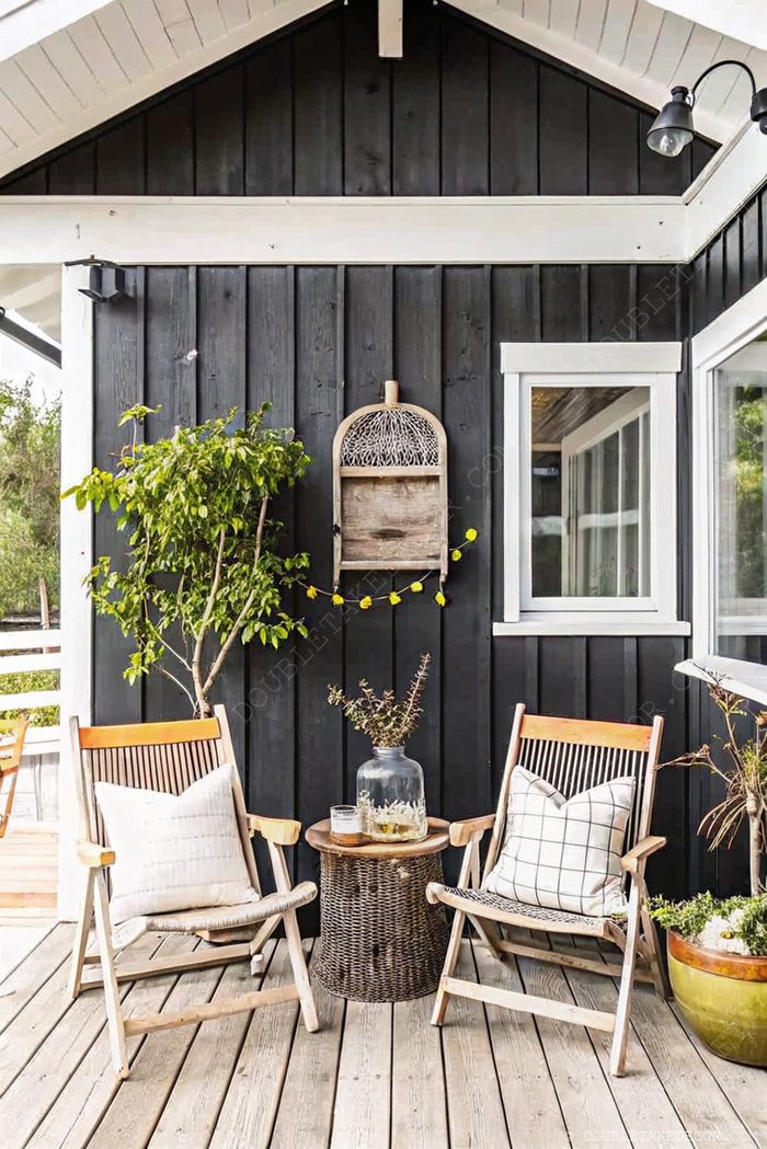 Cozy porch with black wooden wall, white trim, two wooden chairs with cushions, and a woven side table with refreshments.