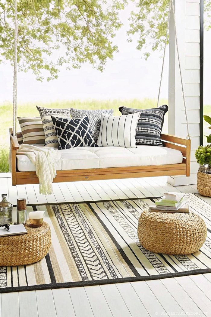 Serene porch with suspended wooden swing bed, plush cushions, and woven poufs on a striped rug, creating a cozy outdoor retreat.