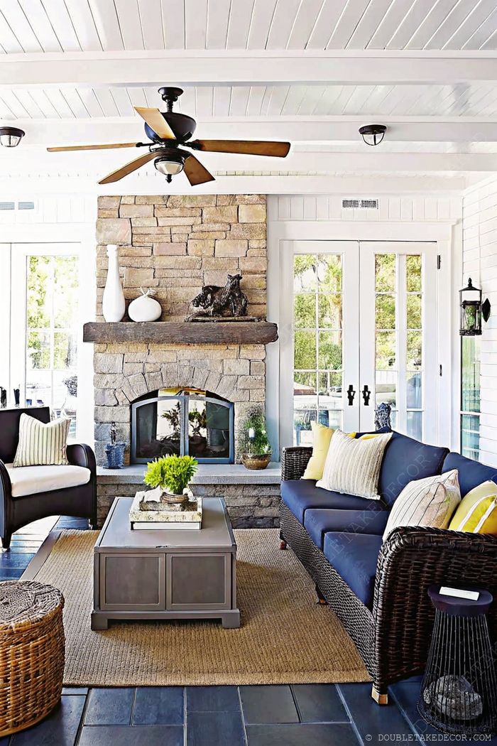 A bright porch with white paneled ceiling, stone fireplace, navy sofa with decorative pillows, and wicker chair creating a comfortable outdoor living space.