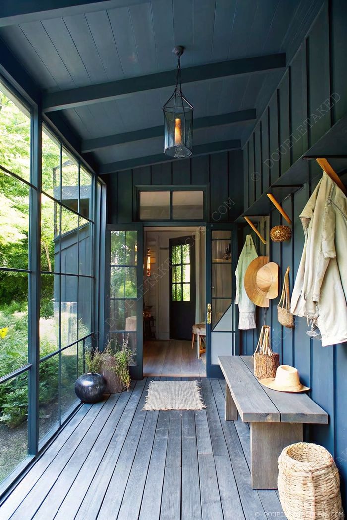 Inviting mudroom with navy walls, wooden bench, hanging hooks, and natural light through glass panels showcasing outdoor greenery.