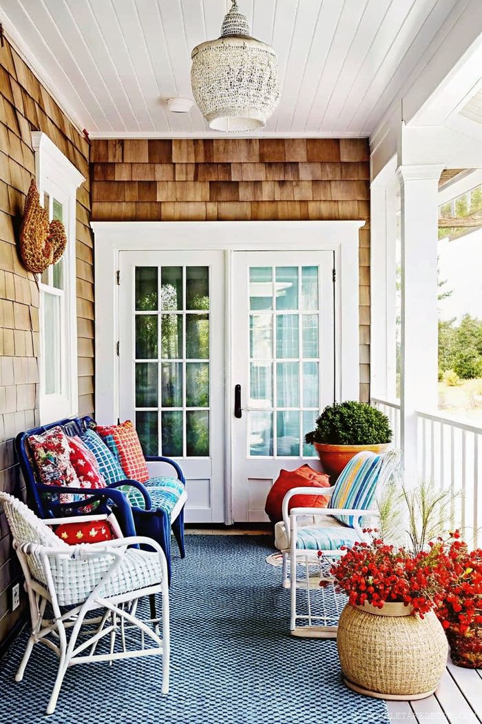 Cozy porch with blue wicker sofa, white chairs, colorful pillows, and potted plants on a woven rug under hanging light fixture.