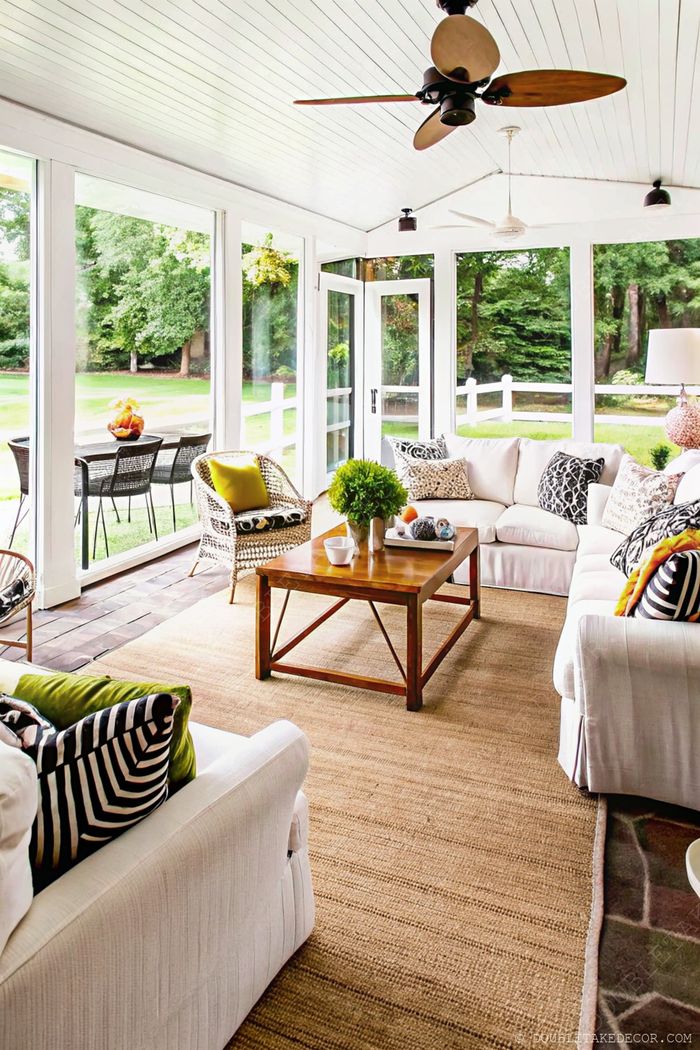 A stylish sunroom featuring white sofas with colorful pillows, woven chairs, wooden coffee table, natural fiber rugs, and large windows overlooking greenery.