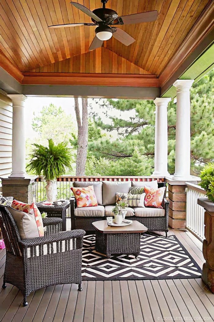 A cozy porch featuring a ceiling fan, plush sofa with colorful pillows, wicker chairs, and geometric rug surrounded by greenery.