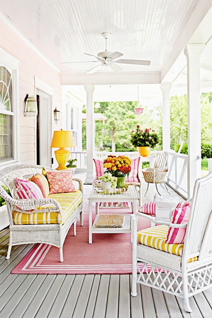 Vibrant porch with white wicker furniture, pink-orange cushions, striped rug, and yellow lamp, surrounded by greenery.