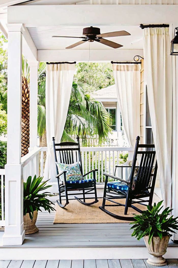 Charming porch with black rocking chairs on a gray deck, featuring blue cushions, white curtains, and tropical plants.