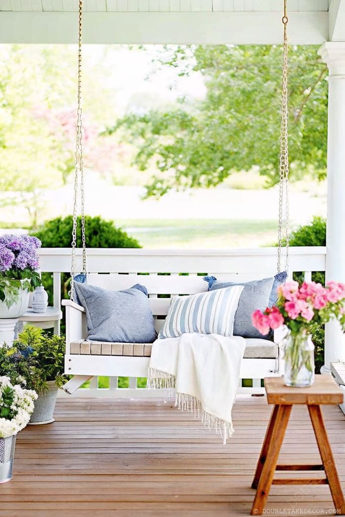 Cozy wooden swing bench with blue and white cushions on a warm wooden deck, surrounded by potted flowers and greenery.