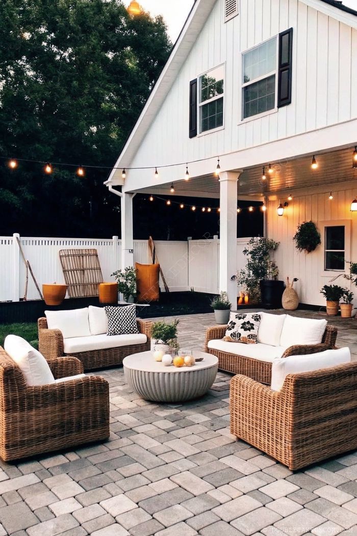 Inviting outdoor living space with wicker furniture, white cushions, stone coffee table, and string lights creating a cozy porch atmosphere.
