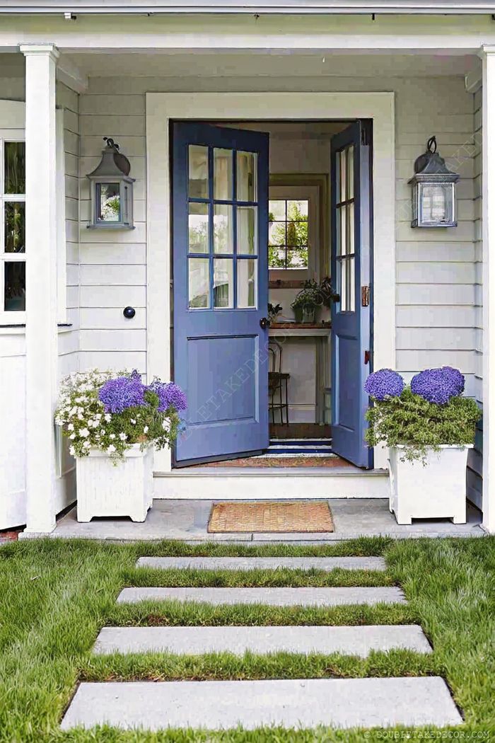 Charming home entrance with blue double doors, white planters with purple flowers, stone pathway and wall-mounted lights