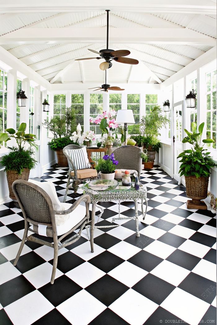 Véranda lumineuse avec sol en damier, fauteuils en osier, table basse en verre et nombreuses plantes en pots baignant dans la lumière naturelle.