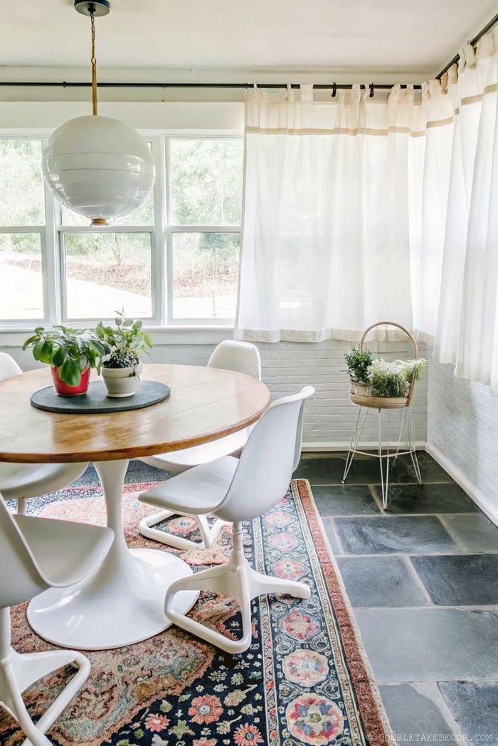 Salle à manger lumineuse avec table ronde en bois, chaises blanches, plantes d’intérieur, rideaux et suspension pour une ambiance cosy de véranda.