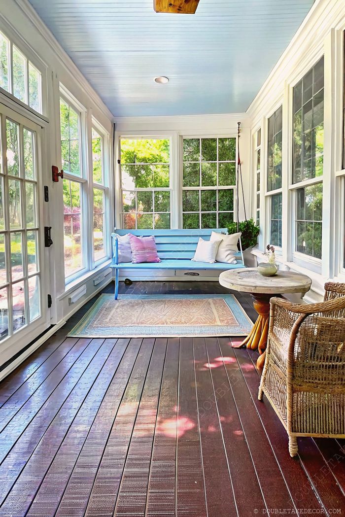 Véranda lumineuse avec plafond bleu, grandes fenêtres, banquette confortable avec coussins, table rustique et fauteuil en osier sur un sol en bois foncé.
