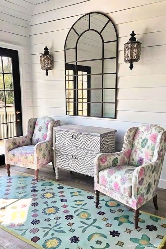 Entrée charmante avec fauteuils fleuris, console géométrique, miroir arqué, appliques vintage et tapis coloré sur murs lambrissés blancs.