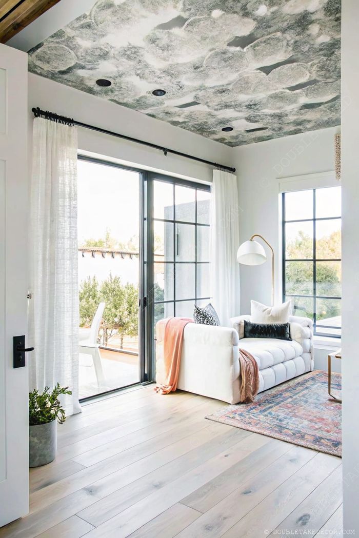 Véranda lumineuse avec grandes fenêtres, canapé blanc et plaid orange, lampe sur pied minimaliste et tapis ancien pour un coin cosy moderne.