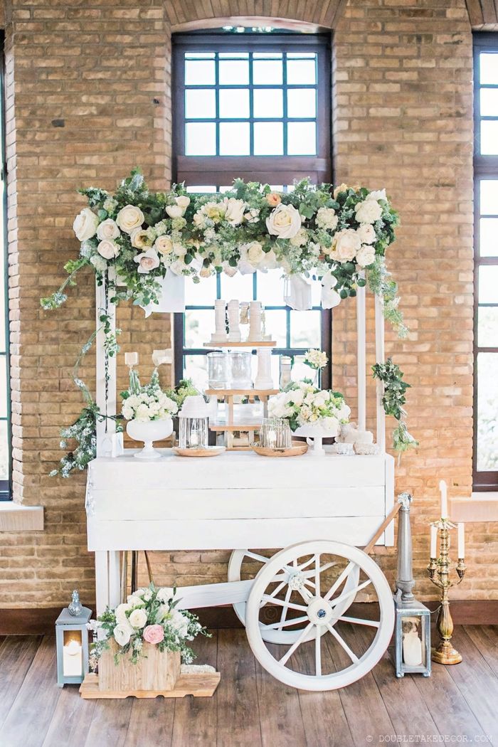 Elegant white dessert cart with white roses and soft flowers against brick walls, perfect for a DIY flower bar at events.