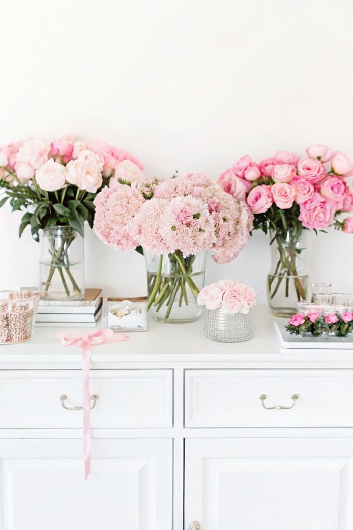 Table with white finish featuring vases of pink roses and chrysanthemums, adorned with books, silver containers, and a delicate pink ribbon.