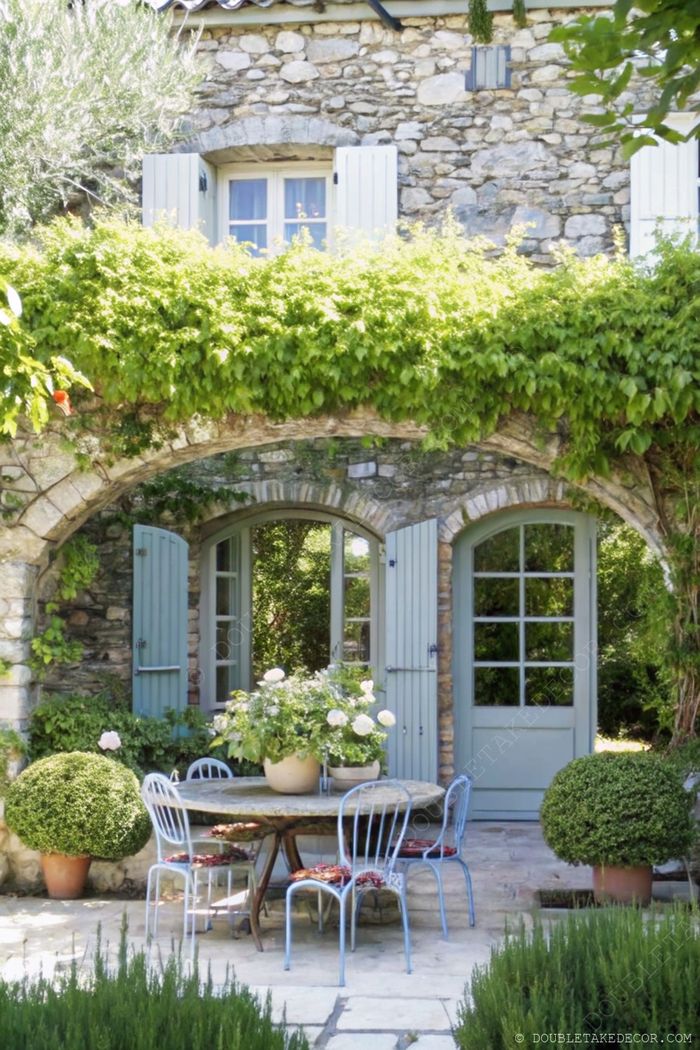 Stone cottage patio with round table, blue chairs, climbing vines, and manicured topiaries creating a serene French garden atmosphere.