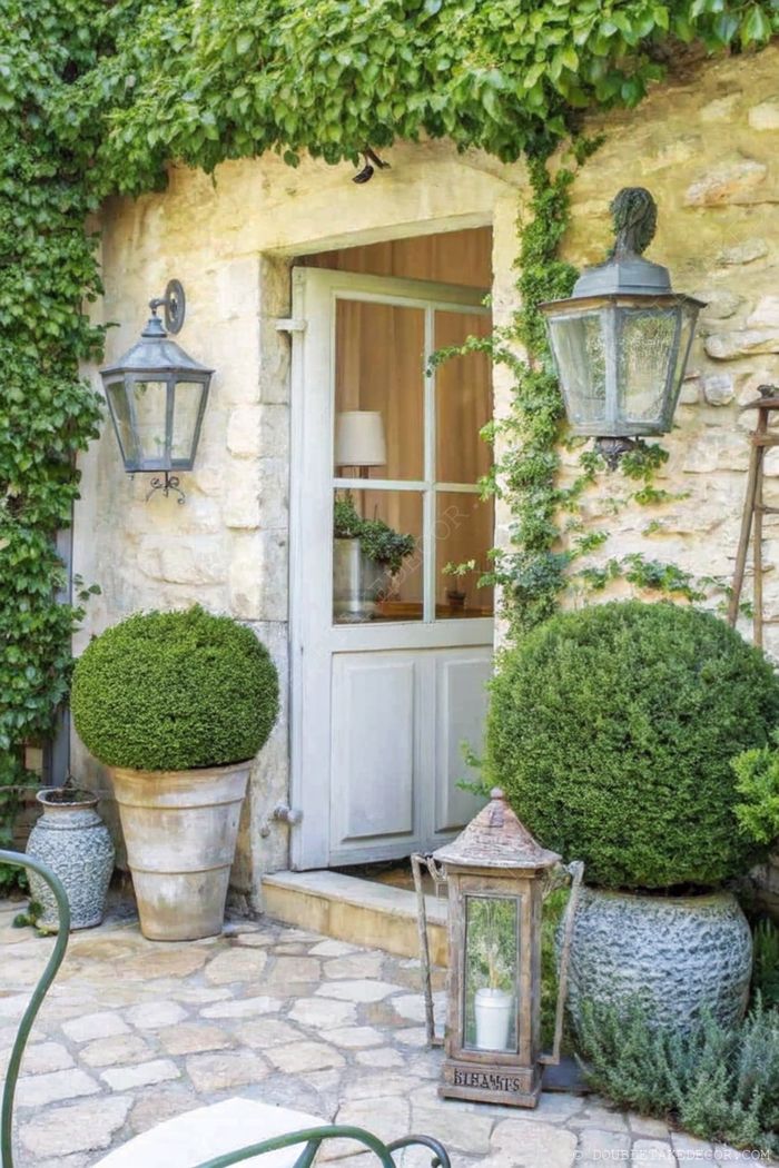 Charming stone cottage entrance with ivy framing, decorative lanterns, and elegant topiary planters along a cobblestone path.