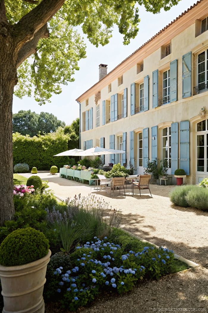 Serene French garden with hydrangeas and lavender bordering a gravel path leading to a yellow home with blue shutters and shaded outdoor dining area.