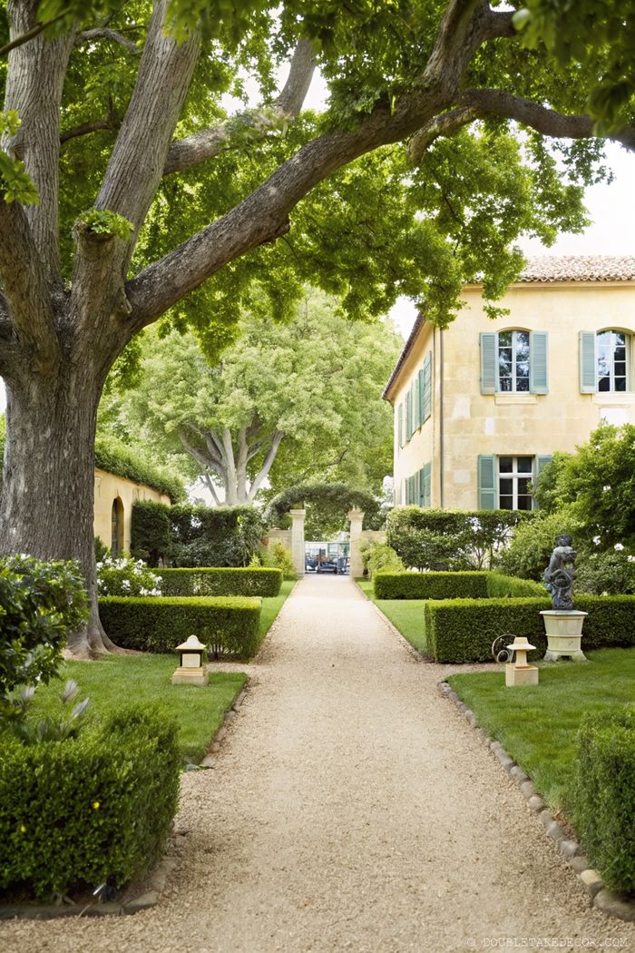 Symmetrical garden pathway with gravel leading to a golden stone French house with green shutters and manicured hedges