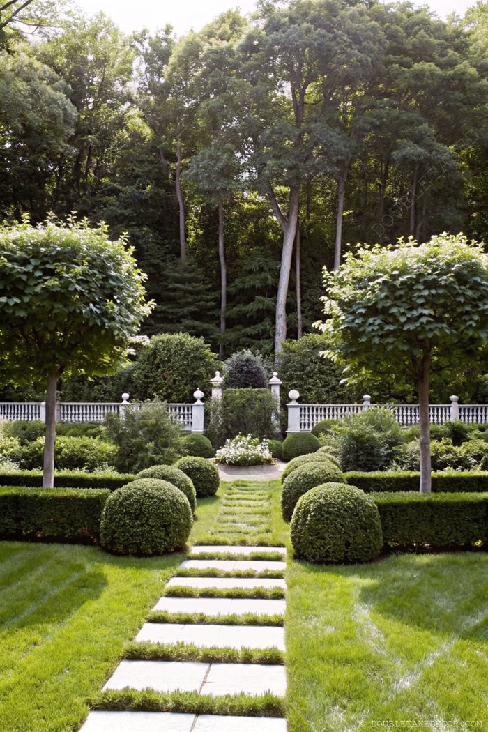 Beautifully manicured garden with stone path through green lawn, bordered by round bushes and trimmed hedges leading to a white decorative fence.