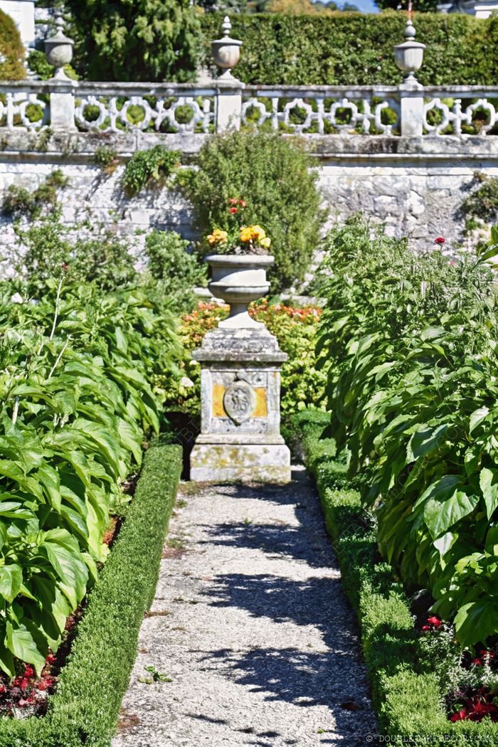 Landscaped French garden pathway with manicured hedges, decorative stone urn centerpiece, and classic stone wall backdrop.
