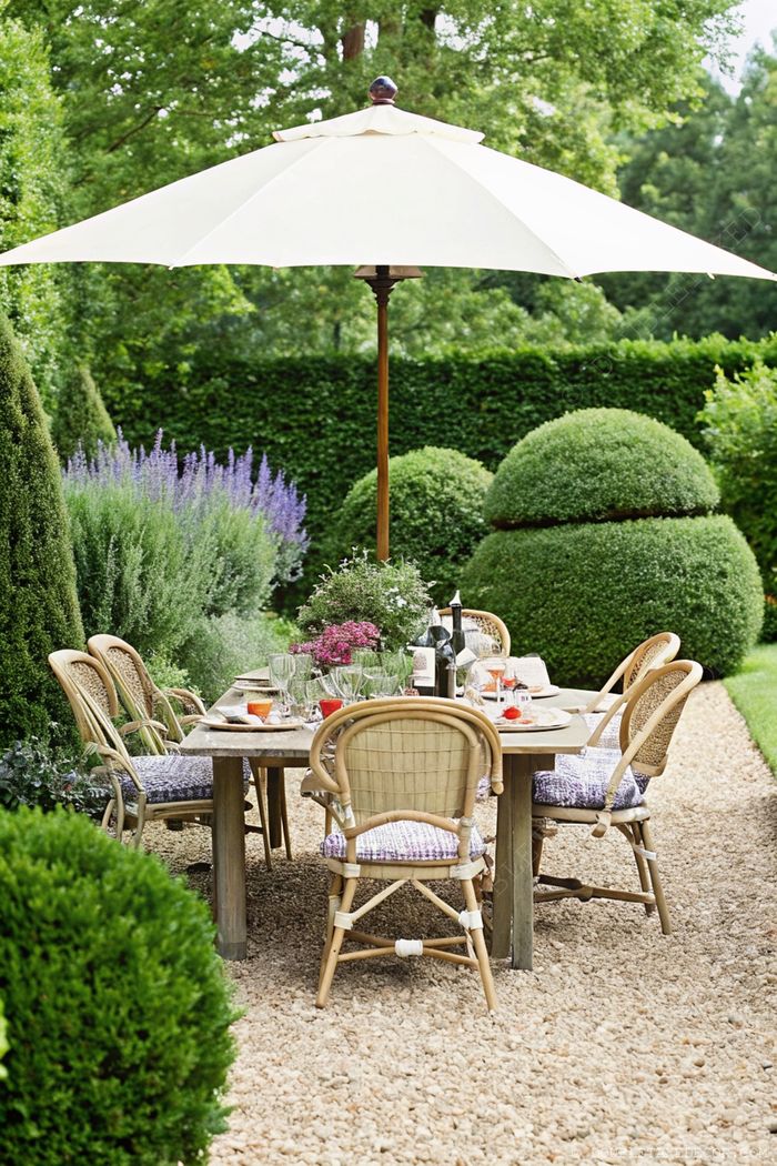 A serene French garden dining area with a cream umbrella, rustic wooden table, wicker chairs, and lavender plants on a gravel pathway.