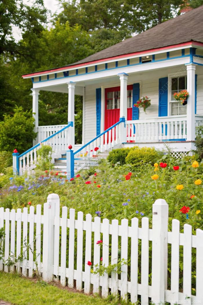 White house with blue shutters and red door, surrounded by vibrant flower gardens and enclosed by a picket fence.