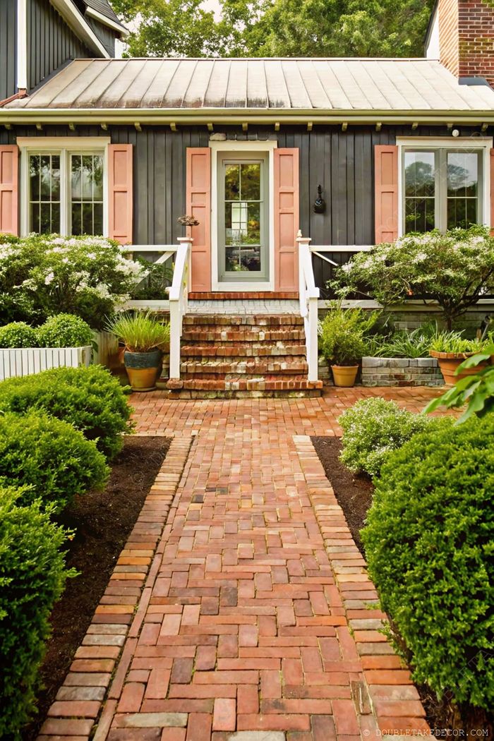 Elegant gray house with pink shutters featuring a brick pathway lined with round bushes and colorful flowers