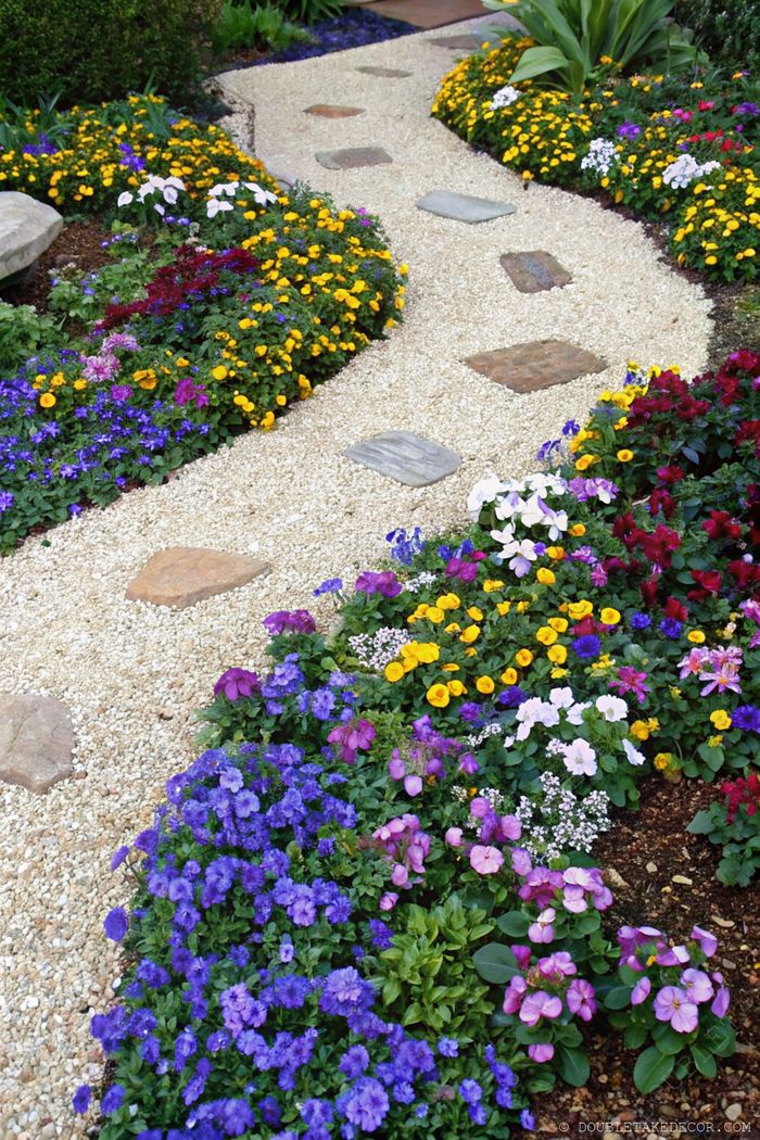 Gravel and stone pathway curving through dense, colorful flower beds with purple, yellow, and red blooms creating a serene garden landscape.