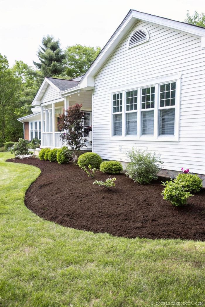Elegant white house with large windows, featuring a curved garden bed with brown mulch, rounded green shrubs, and colorful flowering plants on a well-maintained lawn.