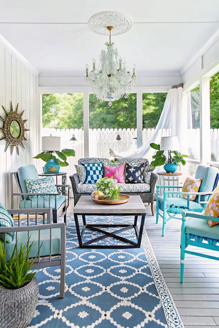 Heller Wintergarten mit grauem Sofa, türkisfarbenen Sesseln, dunklem Tisch, blauem Teppich und üppigen Pflanzen im Sonnenlicht.