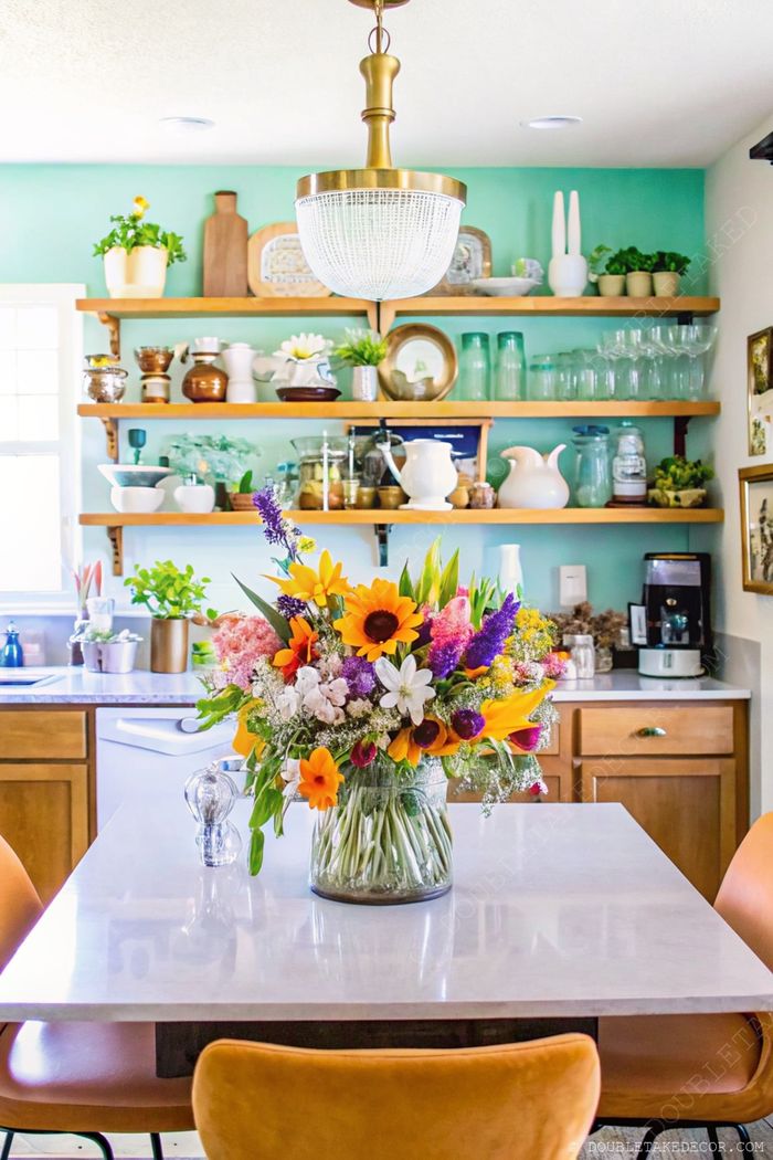 Sunflower bouquet on marble table with open wooden shelves, turquoise walls, and orange chairs creating a warm kitchen atmosphere.