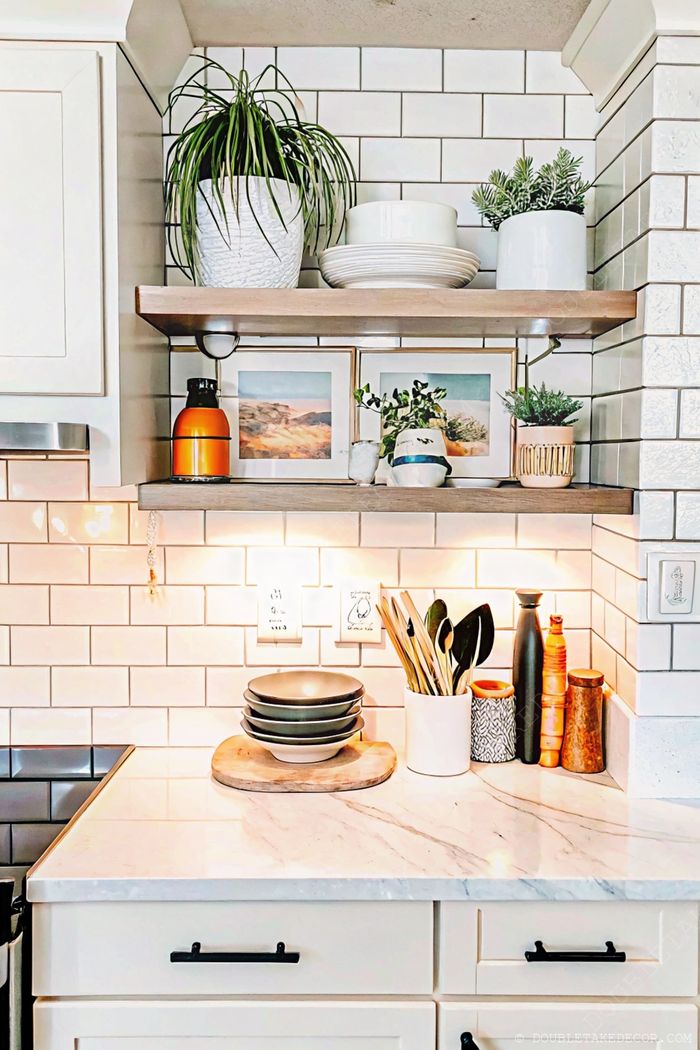 Stylish kitchen featuring wooden open shelves with plants, framed art, and dishware above a marble countertop with organized cooking essentials.