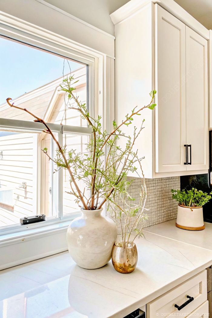 Bright kitchen featuring white ceramic and gold vases with budding branches on a light countertop, complemented by white cabinetry and reflective tile backsplash.