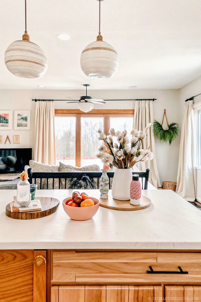 Bright kitchen island with marble countertop featuring a pink fruit bowl, cotton branch centerpiece, and decorative bottles under pendant lighting.