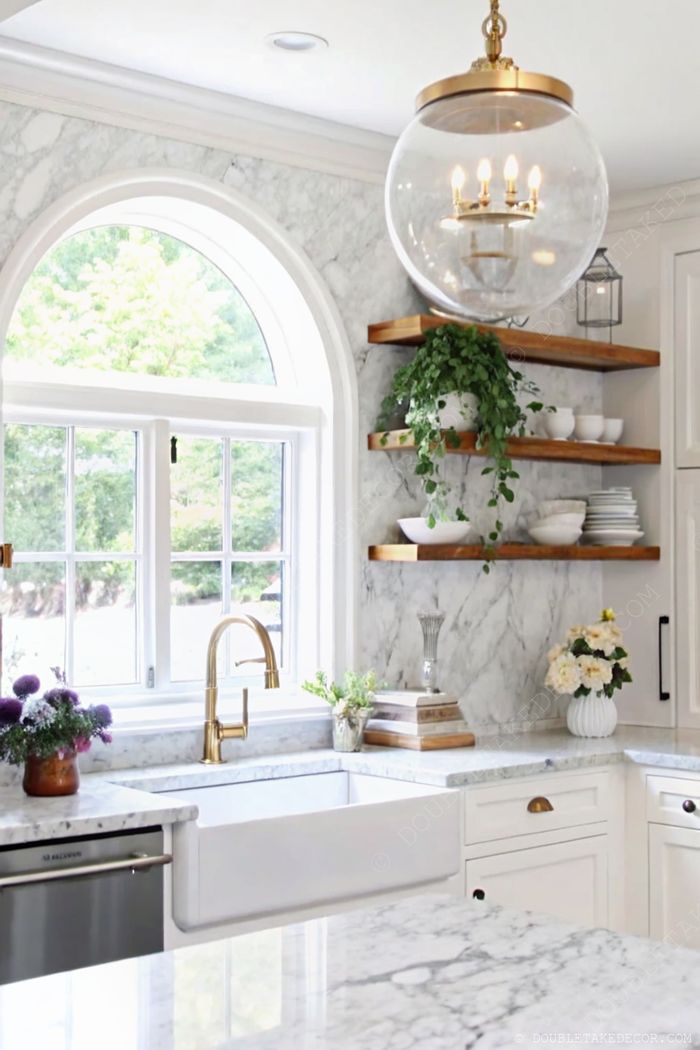 Bright kitchen with marble countertops, gold faucet, arched window, and floating wooden shelves displaying plants and dishware.
