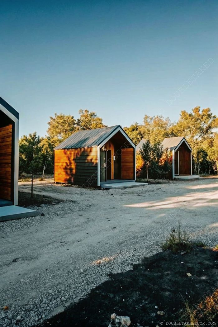 Serene outdoor setting with modern wooden cabins arranged along a gravel path, surrounded by trees and natural greenery.