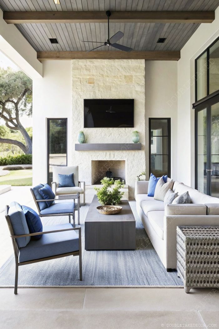 Sophisticated outdoor living space featuring gray sofa, blue armchairs, stone fireplace with TV, and exposed wooden beams.