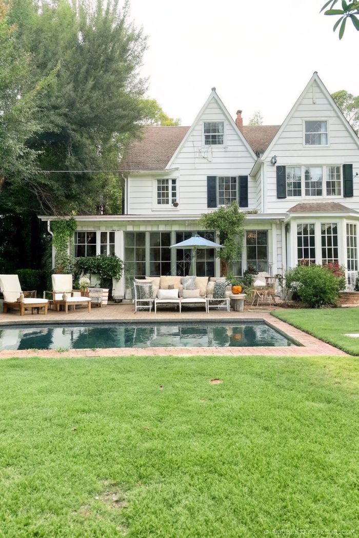 Elegant white house with dark shutters, featuring a rectangular pool, wooden deck with lounge furniture, and shaded seating area surrounded by lush greenery.