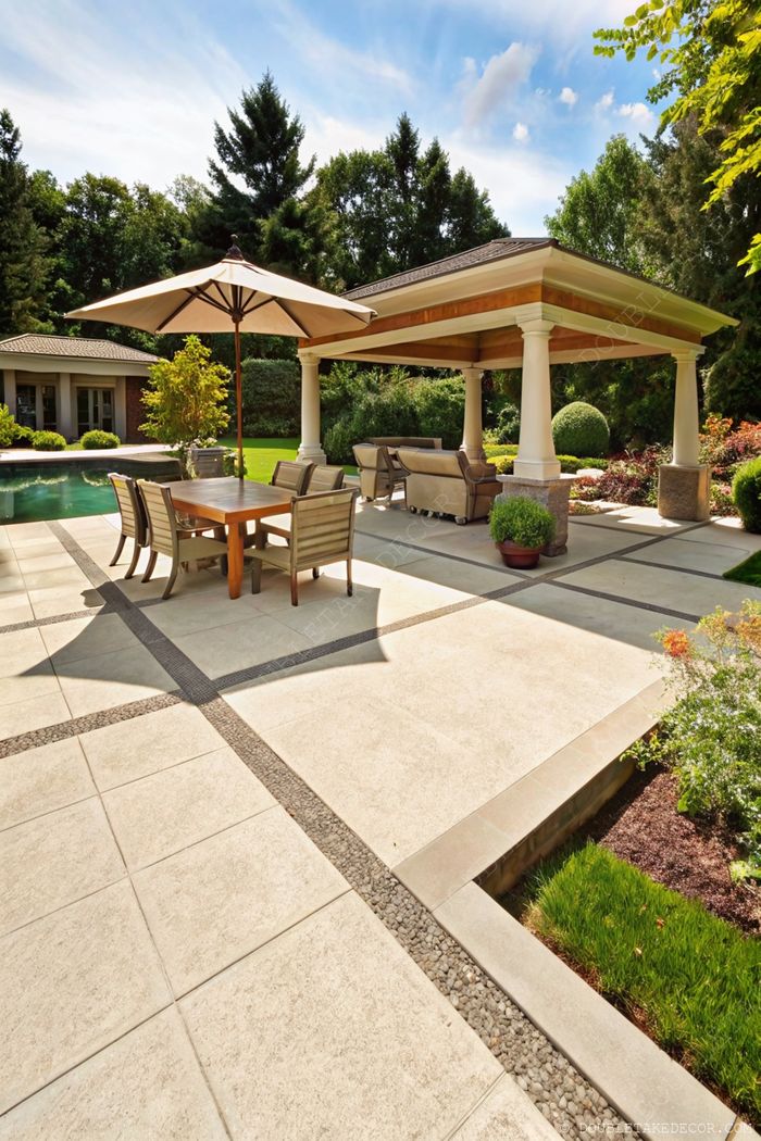 A shaded patio with wooden dining table, modern chairs, decorative stone pathways, covered seating area, and glimpse of pool surrounded by greenery.