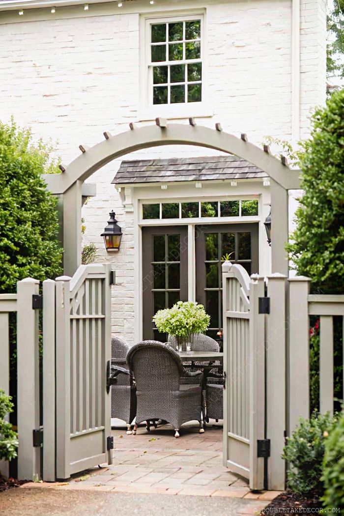 Elegant outdoor dining space with round table, woven chairs, and fresh flowers, framed by a wooden gate and lush hedges.