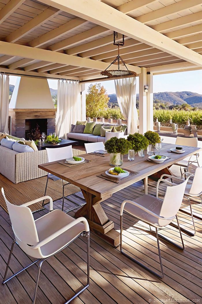 Wooden dining table with white chairs beside a gray sofa with green cushions, framed by white curtains and a stone fireplace.