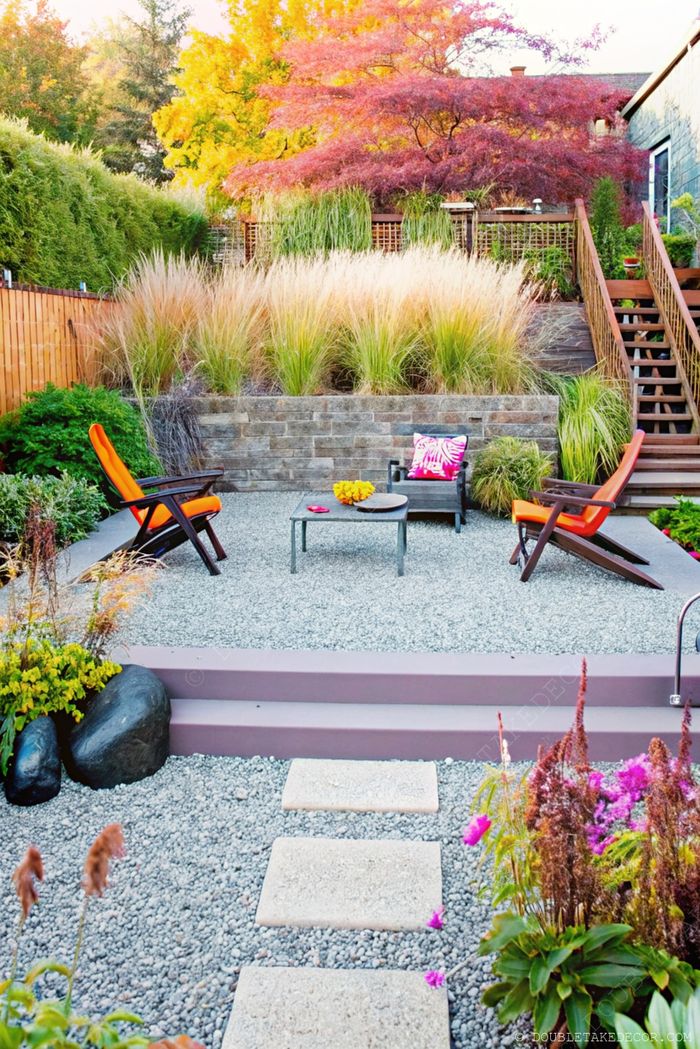 Modern gravel garden with orange and dark lounge chairs around a metal table, surrounded by ornamental grasses and colorful plantings.