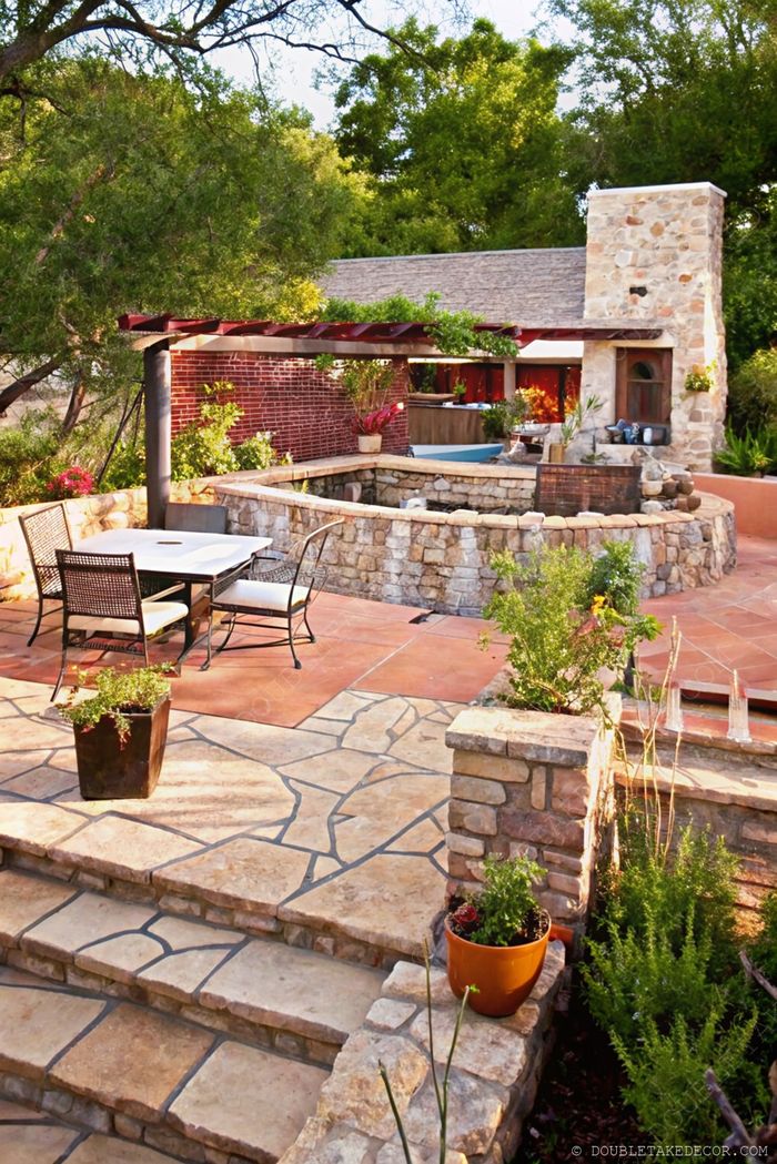 Elegant stone patio featuring a round dining table with black chairs, surrounded by planters and backed by a rustic stone building.