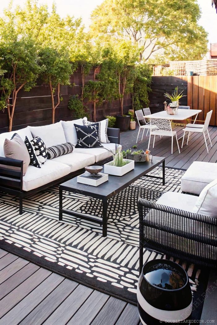 A chic outdoor living space featuring an L-shaped white sofa, black coffee table, patterned rug, and dining area surrounded by greenery.