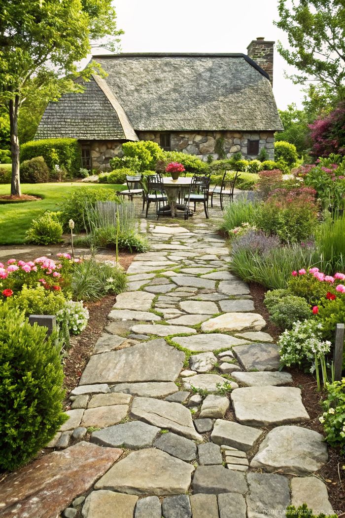 Charming thatched-roof cottage with winding stone pathway through colorful garden, outdoor dining area set on stone patio.
